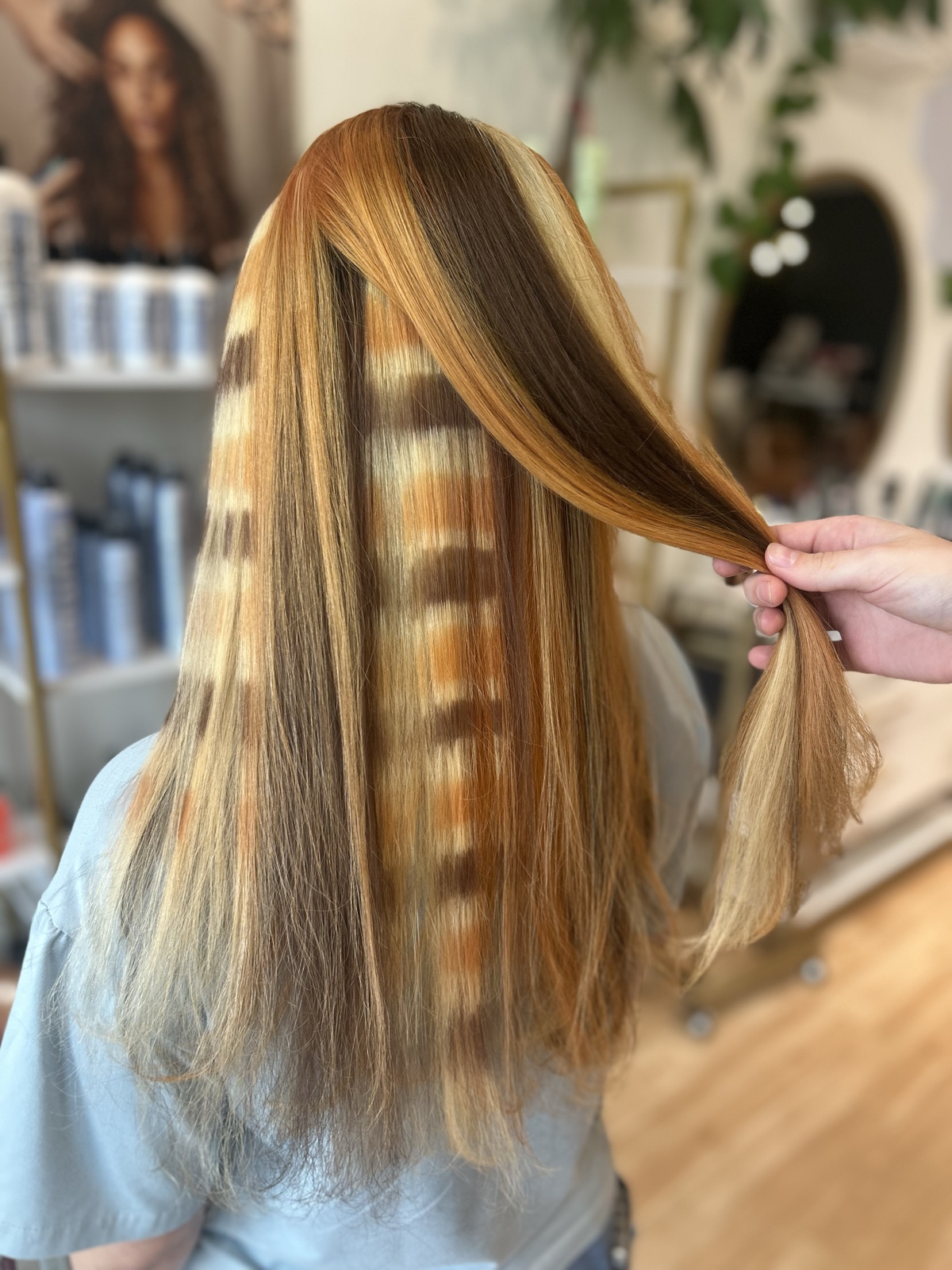 Long straight hair in copper, gold and blonde, with panels banded in brown and copper over pale blonde and one section held aside to show them, seen from behind. Creative color at Canterbury & Co. Salon in Fort Myers