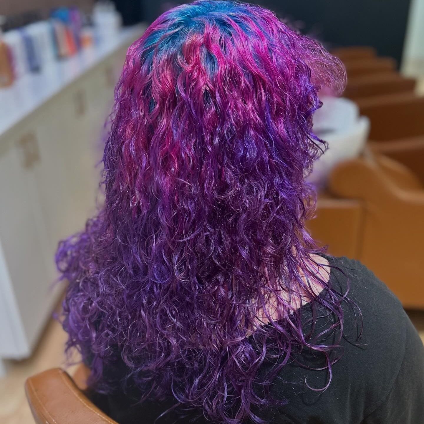 The same curly cut worn down, pink and purple through the curls with blue at the crown, seen from behind